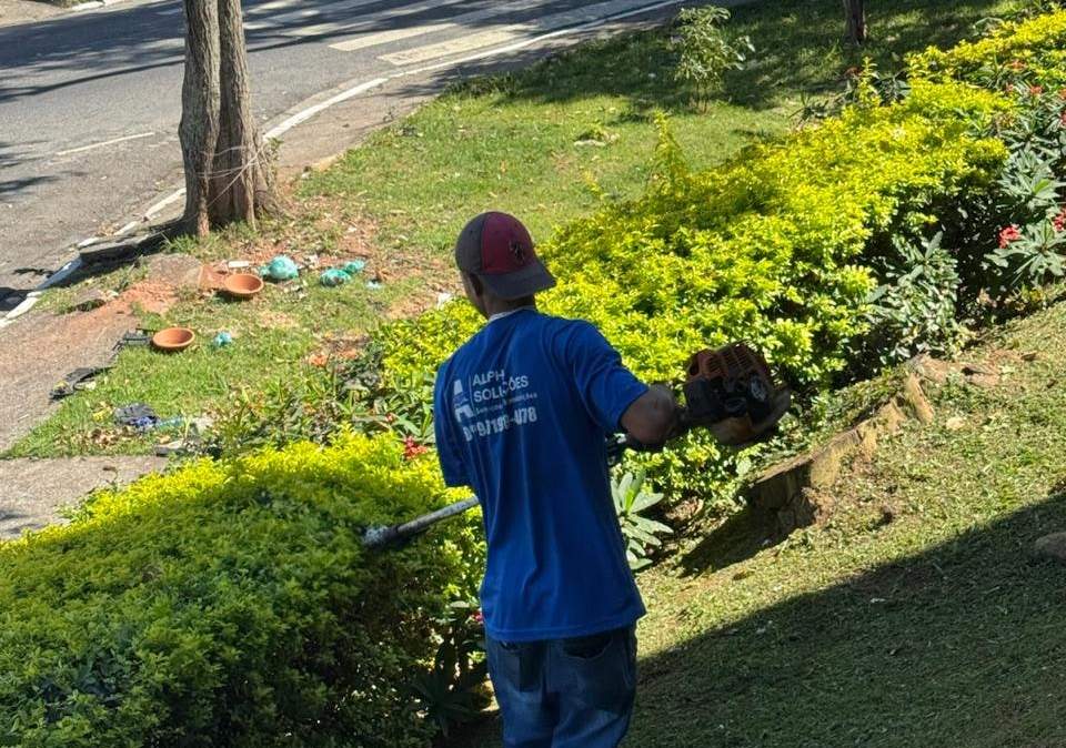 Funcionário da Alpha, de camiseta azul da empresa, aparando um canteiro de arbustos com máquina