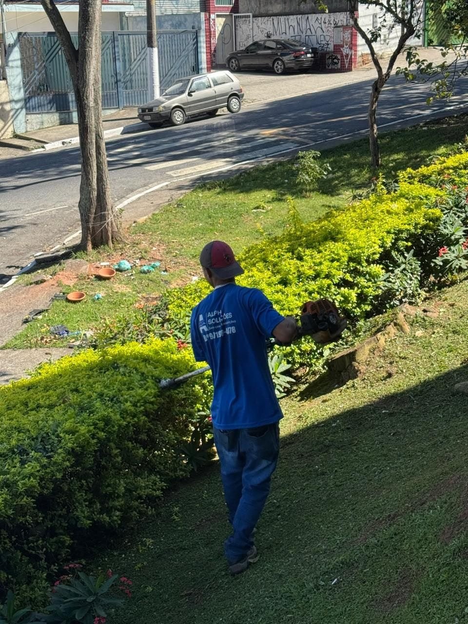 Funcionário da Alpha, de camiseta azul da empresa, aparando um canteiro de arbustos com máquina