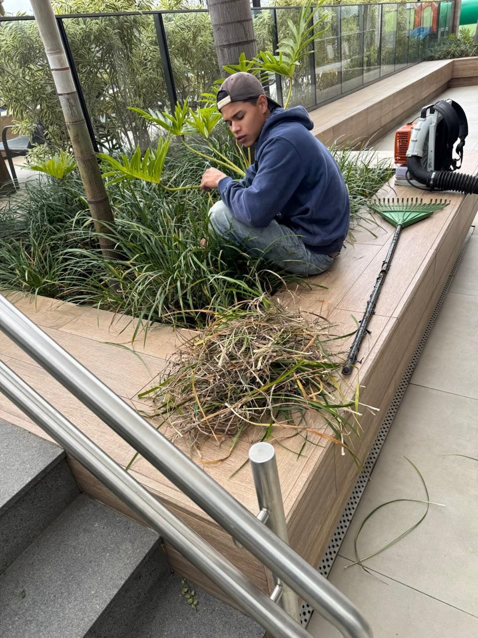 Jardineiro sentado na borda de uma jardineira tirando folhas secas, com o soprador ao lado